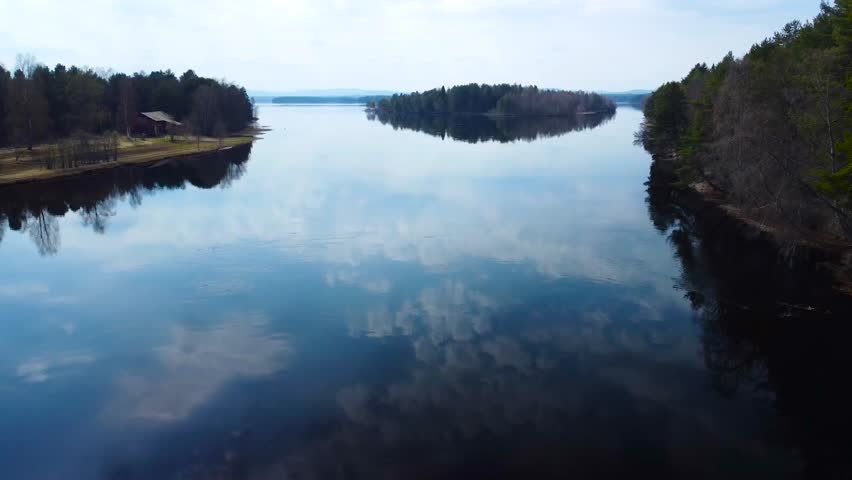 Aerial low flyover over a reflective and glassy blue lake in Sweden during dusk or dawn while the water is reflecting white fluffy clouds and grassy shoreline that have tall pine trees and a cabin.