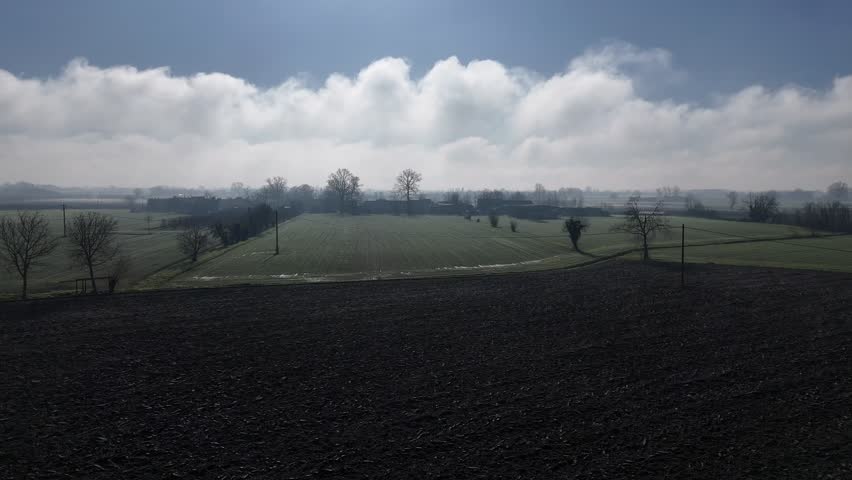 Morning light reveals the rough furrows of a freshly plowed field stretching toward grassy floodplain ridges lined with bare winter trees under drifting clouds above Olza di Castelvetro Piacentino