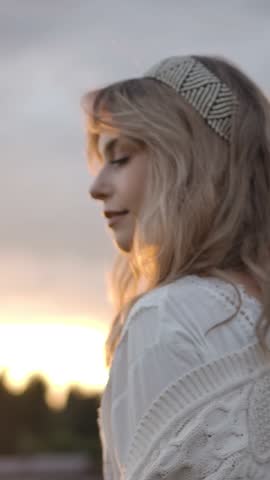 Wonderful blond woman spinning and adjusting blond hair, then looking at camera against overcast sundown sky in countryside. Vertical shot.