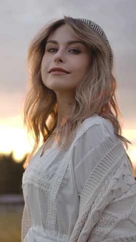 Coquettish lady with stylish headband smiling and touching blond hair while standing in countryside field against overcast sundown sky. Vertical shot.