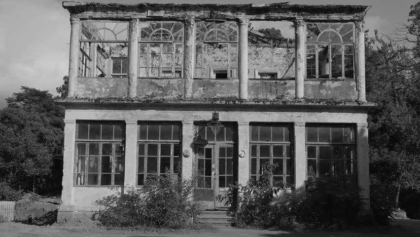 Crumbling facade of a two-story neoclassical building with broken arched windows and weathered pillars, evoking decay and abandonment in monochrome. Documentary shot. History. Background