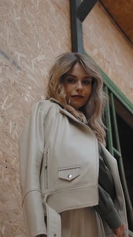Low angle of stylish female smiling and folding arms under jacket, then looking at camera near wall of stable on ranch. Vertical shot.
