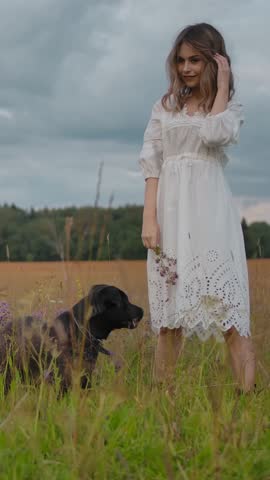 Full body female in white lace dress touching hair and looking away while standing with dog on green meadow against overcast sky in nature. Vertical shot.