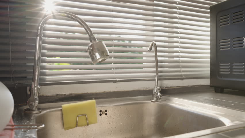 Hand Demonstrating Flexible Faucet by Directing Water Stream in Different Directions Inside a Stainless Steel Sink with Sunlight Through Blinds, Product Showcase Concept