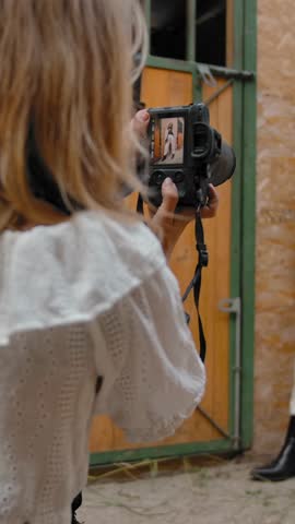Back view of woman sitting on haunches and taking photos of elegant female model douching hair near wall of stable on ranch. Vertical shot.
