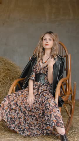 Beautiful sexy girl model, in summer skirts and a leather black jacket, sitting and swinging on a rocking chair near the hay in the barn on the ranch. Vertical shot.