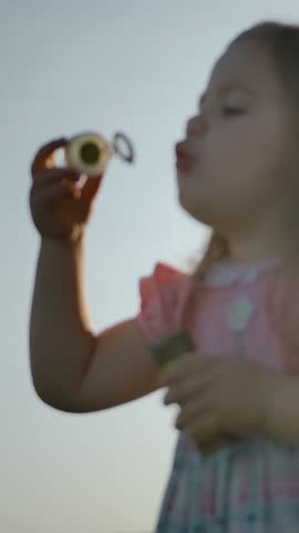 Side view of little girl in blows soap bubbles on the nature. Close-up handheld shot of a happy child. Healthy lifestyle for children in the fresh air outdoor. Far from the city. Slow motion. Vertical