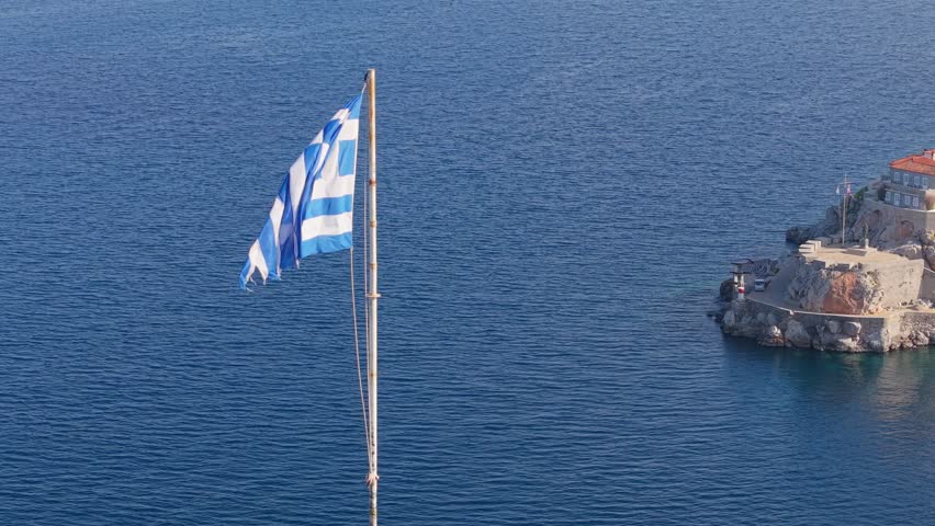 A dynamic drone orbit around the Greek flag atop Hydra’s hill, gradually revealing the historic Miaoulis statue and the island’s traditional harbor below