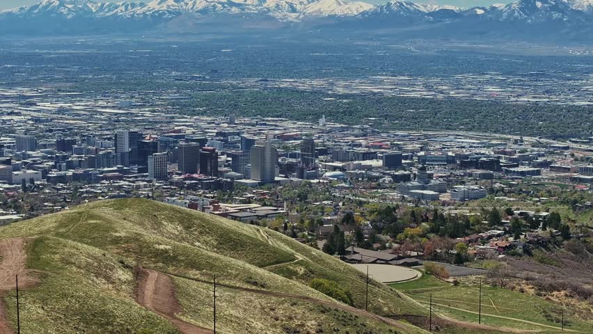 Wide aerial drone shot dollying out and tilting up revealing Salt Lake city, the surrounding valley, and rocky mountains in the background from the hills above on a warm spring day.