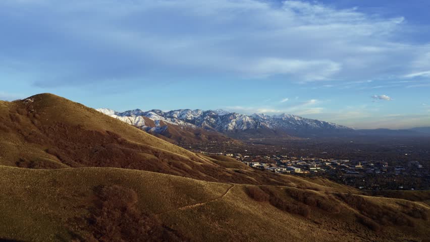 Wide aerial drone shot dollying out over the dry rolling hills of the Bonneville Shoreline trail in Salt Lake City, Utah looking out at the valley surrounded by large snow capped rocky mountains