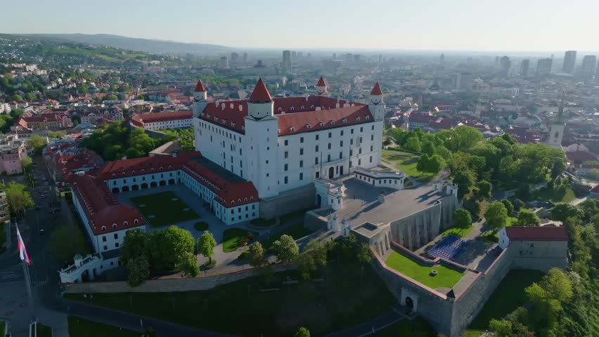 Beautiful drone view of Bratislava Castle bathed in morning light, with the Slovak flag, historical fortifications, and modern city skyline in the background.