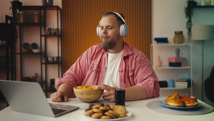 Plus-size man in headset watching video and eating junk food, obesity issue