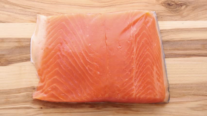 Fresh raw salmon fillet on wooden cutting board being sliced with knife into portions. Chef carefully cuts through the orange flesh creating even pieces for cooking preparation.