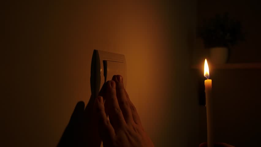 A person reaches for a light switch in low light as a candle flickers nearby during a power outage. Close up
