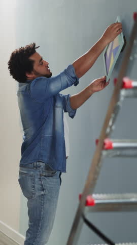Black guy advises brother to turn abstract picture on wall in studio side view. Young African-American designers work in renovated apartment