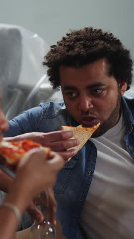 Cheerful African-American man eats pizza slice and talks to friend sitting near foiled sofa closeup. Young builders rest after repairing works in flat