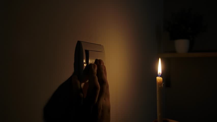 A person reaches for a light switch in low light as a candle flickers nearby during a power outage. Close up