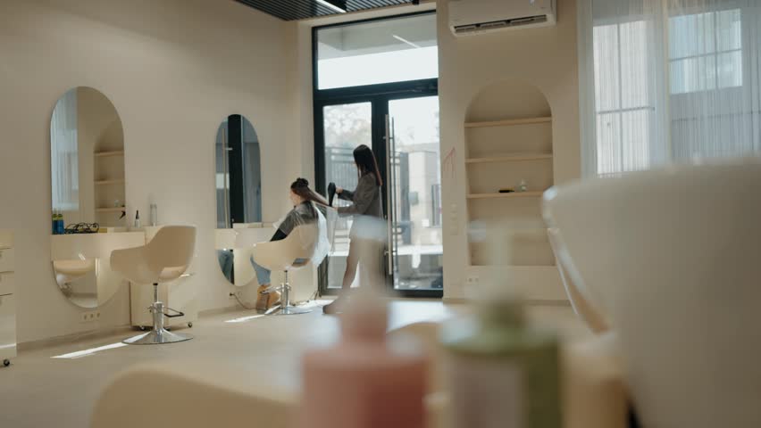 A young woman is getting her hair styled at a modern hair salon. The stylist uses a hairdryer to shape the client's long dark hair. The salon is bright, clean, and minimalist, Modern Beauty Salon