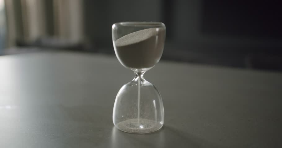 A hand gently flips an hourglass, symbolizing the passage of time. The fine sand flows, creating a visual representation of moments slipping away. This evokes a sense of urgency