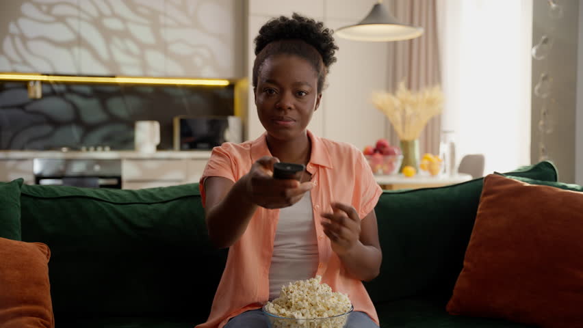 Happy interested African American woman female lady girl businesswoman homeowner eating popcorn turn on TV controller choosing channel watching television film movie series at home weekend chill relax