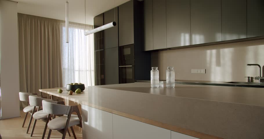 Modern kitchen interior with natural light, featuring an island counter with pears, a dining area, and sleek cabinetry. The minimalist design creates a calm and inviting atmosphere.