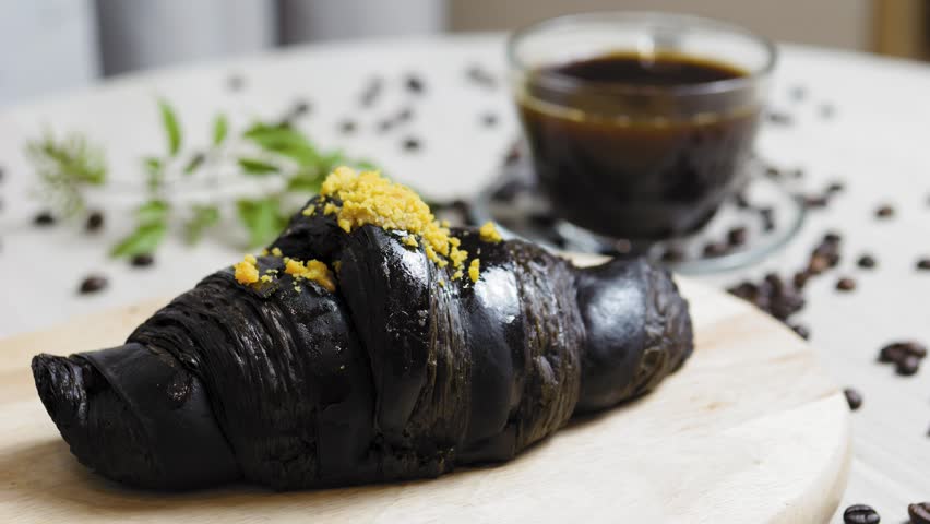 Charcoal croissant sprinkled with yellow crumble beside steaming coffee, resting on wooden surface near scattered coffee beans and green plant leaf