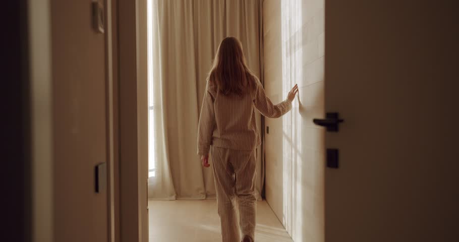 Woman in pajamas walks through dark room into bright bathroom. Cozy home. Morning routine. Getting ready for the day. Comfort and relaxation. Domestic life.