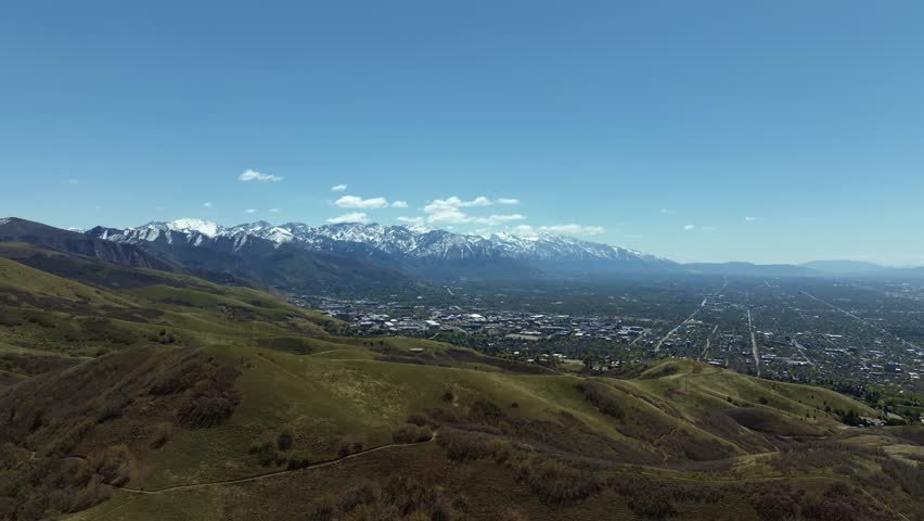 Extreme wide aerial drone shot trucking right from the green rolling hills looking out at the Salt Lake City valley surrounded by large snow capped rocky mountains on a warm sunny spring morning