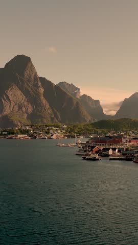 Sunlight bathes the Lofoten Islands in a warm glow. The tranquil waters reflect the rugged mountains, while vibrant structures line the harbor, inviting exploration and adventure, Reine, Norway