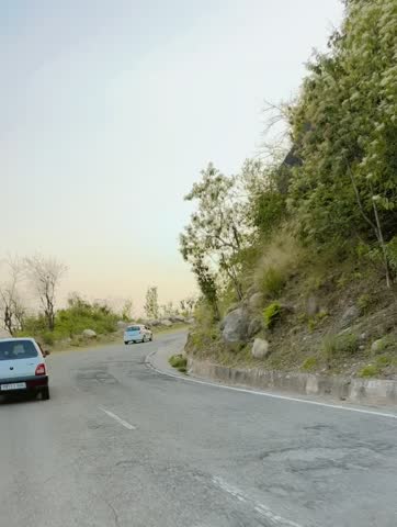 Scenic drive through a winding hill road during sunset, surrounded by lush greenery and trees. Vehicles moving along the curved road with a peaceful and serene natural landscape.