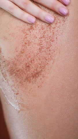 Young slim woman puts anti-cellulite coffee scrub on soft skin of buttocks and thigh in shower at home against blurred wall slow motion closeup