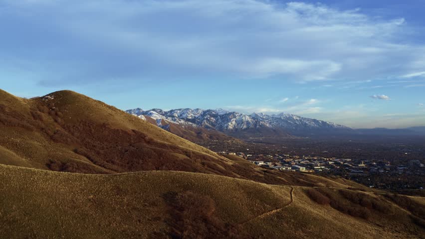 Wide aerial drone shot trucking right over the dry rolling hills of the Bonneville Shoreline trail in Salt Lake City, Utah looking out at the valley surrounded by large snow capped rocky mountains