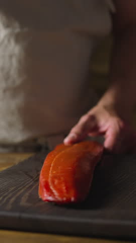 Chef in apron chops delicatessen cool smoked chum salmon fillet with knife on wooden board at table closeup slow motion. Cooking dinner of seafood