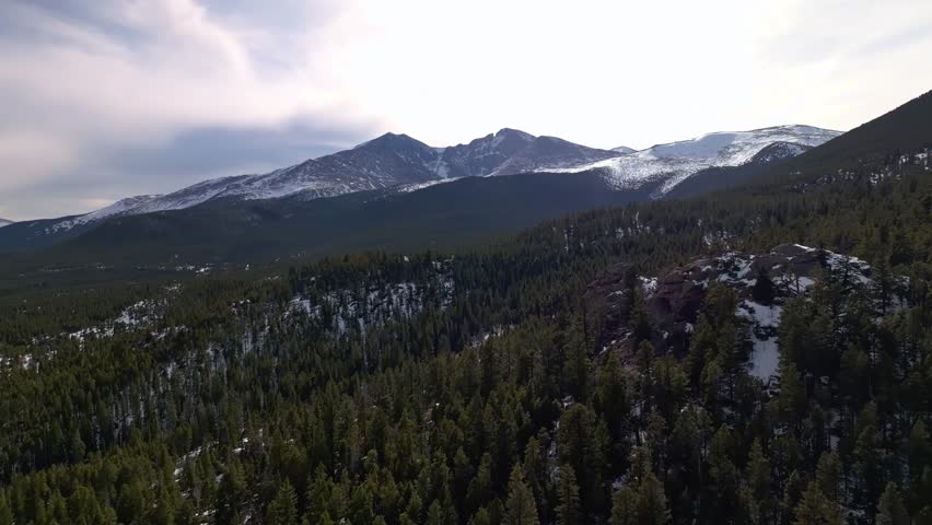 Aerial pullback over rocky pine hillsides with snowy peaks and wide blue sky in the distance, Allenspark Colorado USA