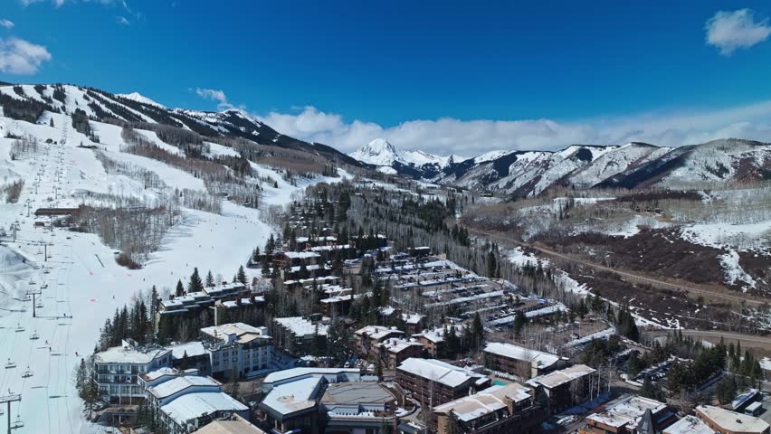 Expansive view over ski runs lined with trees, snowy terrain and mountain peaks beyond, ski in ski out condos and parking lot, Snowmass Colorado