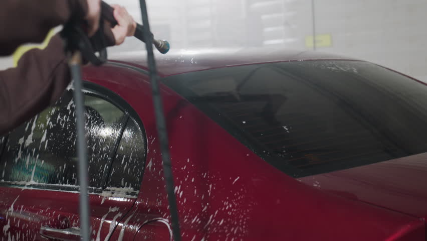 Rear view of person using high pressure hose to rinse soap foam from back of red car inside professional garage environment with water streams and suds flowing dynamically over vehicle surface