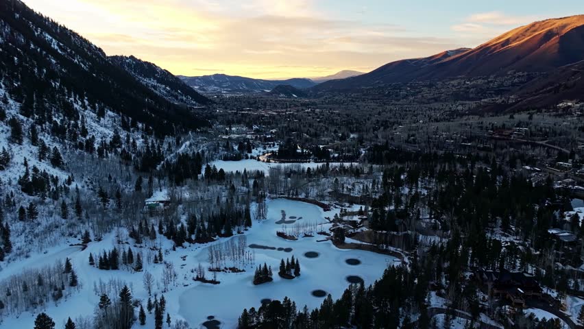 Drone descends above icy river gorge lined with winter trees, shadows stretch across snowbanks, stunning golden hour sunset glow in sky