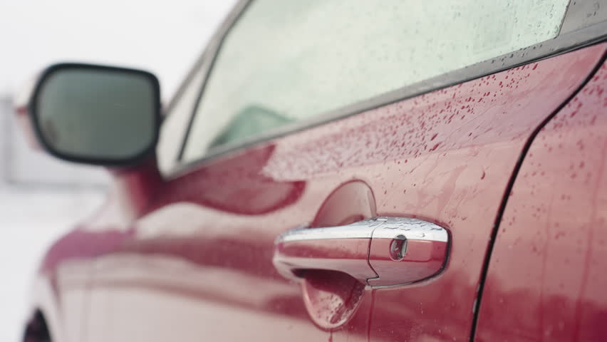 Man wearing sunglasses steps out of red car during light snowfall with visible snow-covered background, vehicle interior in view, and water droplets on car door