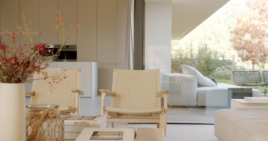 A modern, minimalist living room with two wicker chairs and a coffee table. Large glass doors open to a patio with a white sofa. The kitchen with white cabinets is visible in the background.