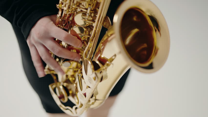 A young woman in black dress plays the saxophone on the white background in studio. Romantic music. Young female musician playing a saxophone. Jazz musician playing the saxophone.