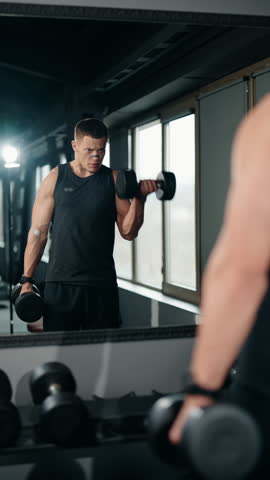A muscular man lifts dumbbells in front of a mirror at the gym. He is focused and determined as he works on his biceps. He is working out hard and looking to grow his muscles.