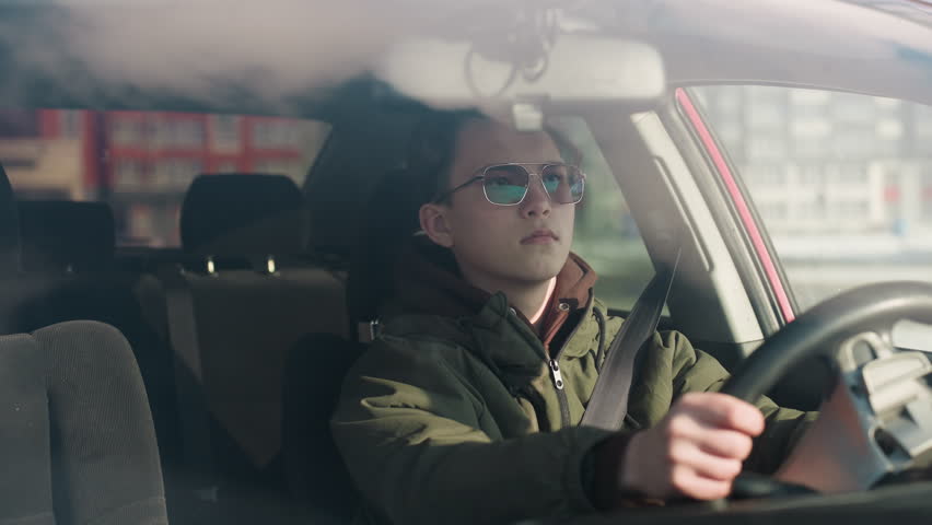 cab driver in parked winter vehicle unfastens seatbelt and grips steering wheel while focusing on device inside cabin with soft daylight and blurred urban background visible through window