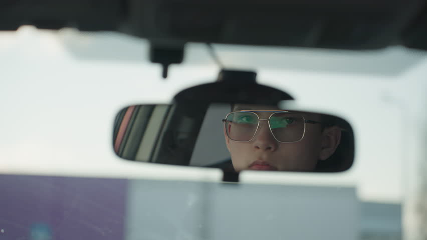rear view mirror reflecting young driver wearing glasses adjusting mirror upward with hand inside parked car with soft focus winter background and diffused daylight through windshield