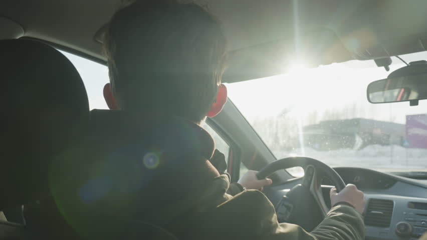 rear view of man dancing in car cabin while steering wheel held, sunlight filtering through windshield onto snow covered parking lot with blurred buildings