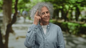 Elderly woman with grey hair gesturing confidently in a sunlit park, wearing a casual denim shirt, surrounded by lush greenery and trees on a peaceful day outdoors. - Powered by Shutterstock - Get 15% off with code: PIKWIZARD15