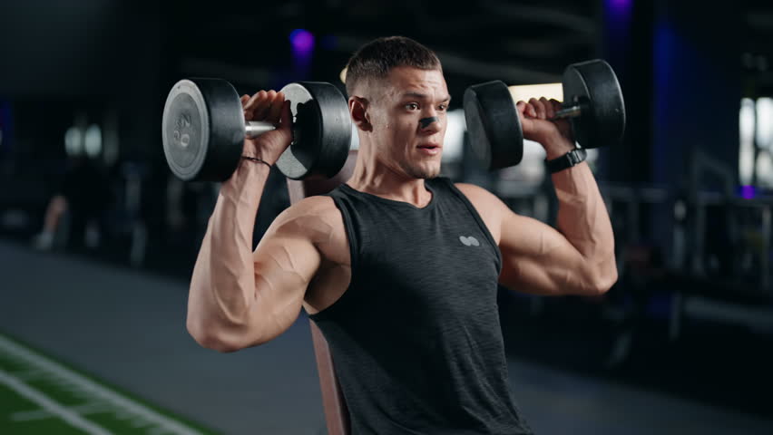 A determined male athlete performs dumbbell shoulder presses in a modern gym. He focuses on proper form and strength training. This video embodies fitness, health, and dedication to exercise.