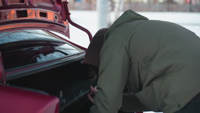 Side view of person in green winter jacket reaching into car boot to remove jack tool, trunk lid open, snowy ground and soft natural lighting in background