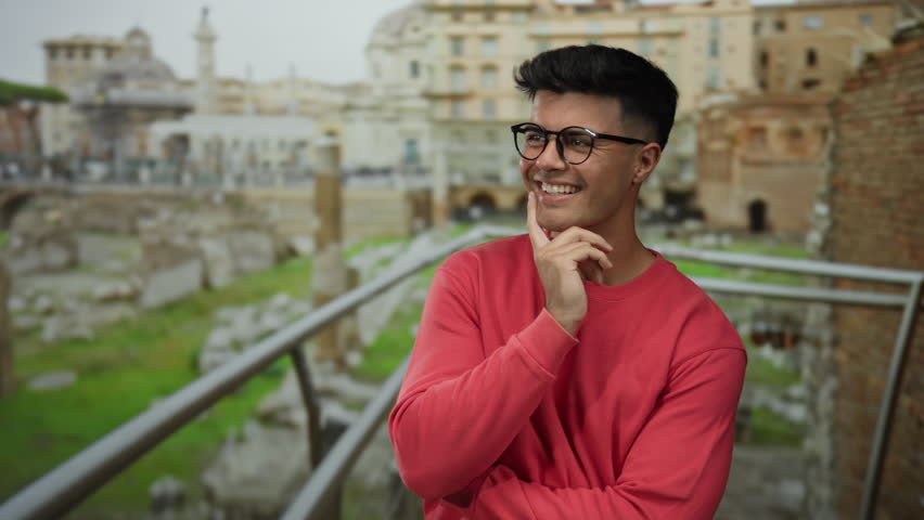 Young man wearing glasses stands thoughtfully at ancient roman ruins in the city, exuding charm and attractiveness in a vibrant red sweater under an overcast sky.