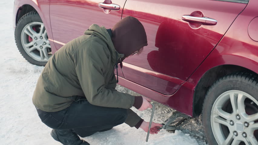 man squatting in winter jacket uses hand to lower jack under car, spanner in hand, snowy ground, car repair, tire change, outdoor action, mechanic work, winter setting, focus on tool use