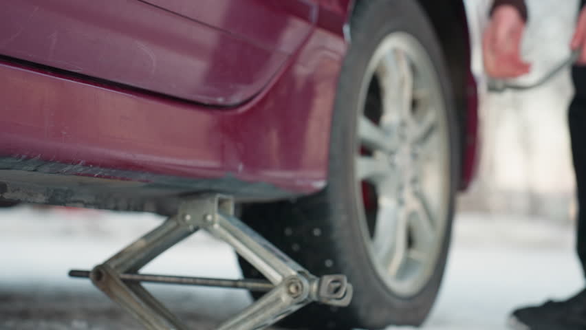 Close up of car jack positioned under vehicle on snowy ground, person slightly loosening bolt on rim, clear focus on tool movement and tire, cold outdoor setting with focus on mechanical work